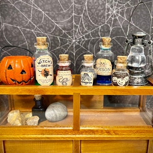 May include: A wooden display case with a glass front contains a collection of miniature glass bottles filled with colorful liquids. The bottles are labeled with Halloween-themed names like "Witch's Brew", "Poison", and "Pumpkin Ale". A small jack-o'-lantern sits on the shelf below the bottles.