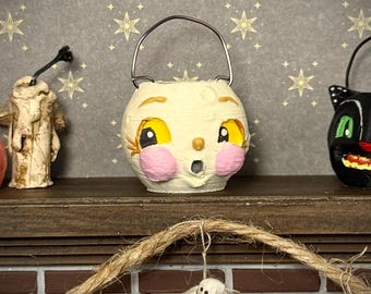 May include: A cream-colored, spherical bucket featuring a painted face with large yellow eyes, pink circular cheeks, and a small open mouth. The bucket has a thin, arched metal handle attached to the top, designed for carrying or hanging.