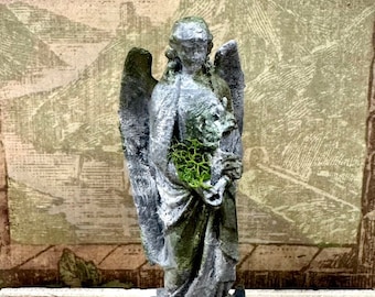 May include: A gray, weathered-look angel figurine standing on a rectangular base. The figure has large wings, draped robes, and holds a cluster of bright green preserved moss in its hands. Additional green moss is attached to the base of the statue.