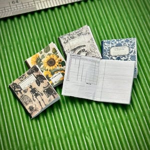 May include: Four miniature notebooks with different designs. One notebook has a sunflower and floral design, another has a skeleton and anatomical illustration, a third has a fish and floral design, and the last has a blue and white patterned cover with the text "Composition Notebook".