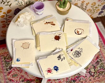 May include: A collection of miniature manila file folders arranged on a white round table. The folders feature various hand-drawn doodles, including a rocket, lightning bolts, scribbles, hearts, and brown coffee-like stains on the paper surfaces.