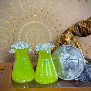 May include: Two small green glass beakers with clear glass stoppers decorated with iridescent beads. The beakers are sitting on a wooden surface.