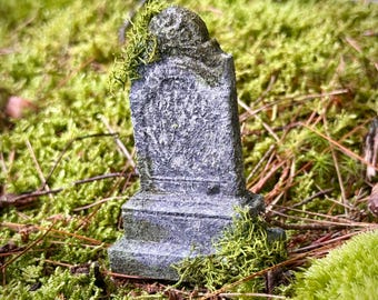 May include: A miniature gray tombstone with a weathered, textured surface and a rounded top. The small monument features a rectangular base and is partially covered with patches of bright green moss, giving it an aged and weathered appearance.