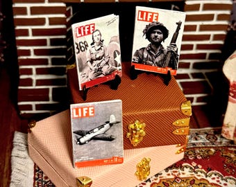 May include: Miniature vintage LIFE magazines with red and white covers, featuring images of a woman, a soldier, and an airplane. The magazines are displayed on a brown trunk and a pink box with gold accents, set against a brick fireplace.