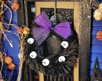 May include: A miniature black wreath featuring four white eyeballs with black pupils attached to the textured surface. A purple mesh bow is secured to the top of the circular wreath, which is suspended against a dark wooden door frame with orange floral accents.