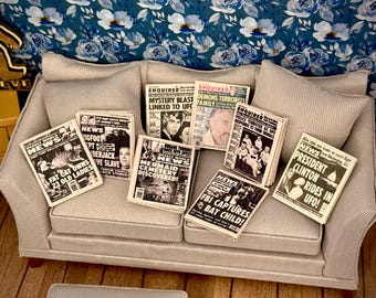 May include: Miniature sofa with several tabloid newspapers scattered on top. Headlines include "Fat Cat Owns 23 Old Ladies," "Mystery Blast Linked to UFC," and "President Clinton Rides in UFO!" The background features a blue floral wallpaper.