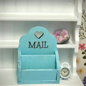 May include: A light blue wooden mail holder with a heart cutout and the word "MAIL" in black lettering. The holder has two slots for mail and is sitting on a white shelf.