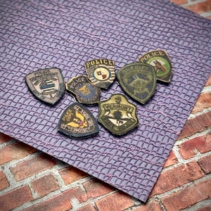 May include: A collection of seven vintage police patches, featuring various designs and colors. The patches are made of fabric and have a worn, distressed look. Some of the patches feature the word "Police" and others feature the names of different police departments.