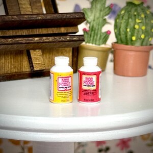 May include: Two miniature plastic bottles of Mod Podge adhesive. One bottle features a yellow label with black text, and the other bottle features a red label with black text. Both bottles have white screw-on caps and are cylindrical in shape.