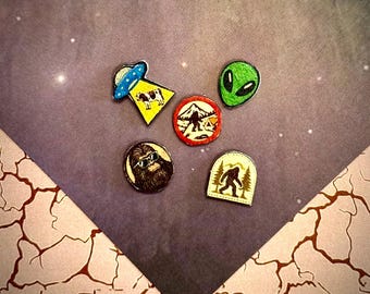 May include: A set of five small pins featuring various designs: a blue UFO abducting a cow, a green alien head, a circular red mountain scene with a creature, a round pin of a furry creature wearing sunglasses, and a beige arch-shaped pin of a creature.