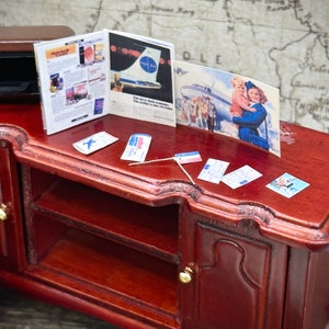 May include: A miniature wooden cabinet with two doors and a shelf. The cabinet is decorated with gold knobs. On top of the cabinet are miniature travel items, including a magazine, a postcard, and several airline tickets. The magazine has a blue and white airplane on the cover and the text "STELLAR" on the front. The postcard shows a vintage airplane with a flight attendant holding a child. The text "DLE" is visible on the postcard. The airline tickets are blue and white with the text "PAN AM" on them.