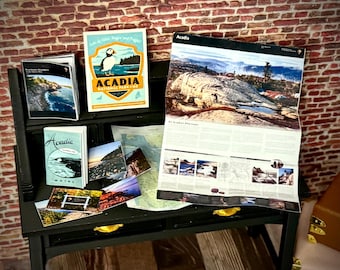 May include: A black miniature desk displays travel brochures and a map. The brochures feature images and text related to Acadia National Park in Maine. One brochure has a puffin illustration and the words "Acadia National Park Maine".