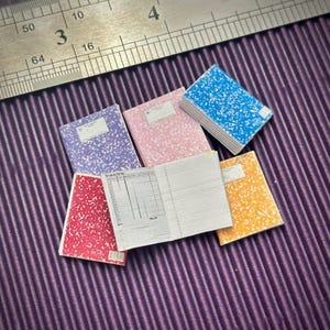 May include: Five miniature composition notebooks with different colored covers. The covers are red, pink, blue, purple, and yellow. The notebooks are on a purple striped surface.