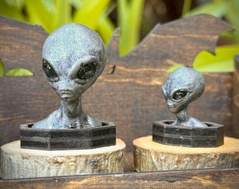 May include: Two silver-toned alien busts with large black eyes, each mounted on a black hexagonal base and a natural light-colored wood slice. The larger alien figure is positioned on the left, while a smaller version is placed to the right.
