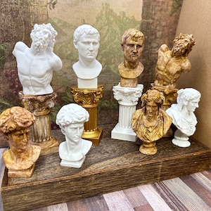 May include: A collection of 10 white and brown plaster statues of various figures, including a man with a beard, a woman with long hair, and a man with a crown. The statues are displayed on wooden stands with gold accents.