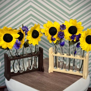 May include: Two miniature floral arrangements featuring yellow sunflowers and purple flowers in glass test tubes. The test tubes are held in wooden racks, one dark brown and one light beige. The background is a light green and white chevron pattern.