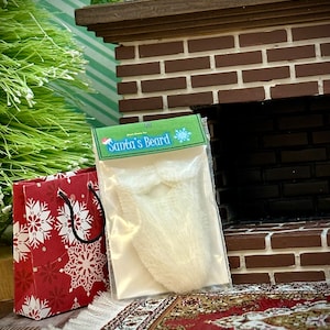 May include: A white faux fur Santa beard in a clear plastic package with a red and white snowflake patterned gift bag and a small house figurine on a brick fireplace mantel.