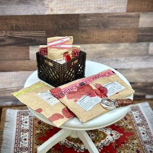 May include: A set of brown paper envelopes with red evidence tape and bloodstains. The envelopes are labeled "EVIDENCE" and have a chain of custody label. The envelopes are sitting on a white table with a red and cream patterned rug. A small, round, silver object is also on the table.
