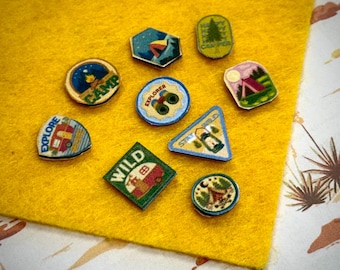 May include: A collection of nine small, flat, decorative pins featuring various outdoor and camping-themed illustrations. The pins include shapes like circles, triangles, squares, and hexagons, displaying colorful graphics of tents, lanterns, trees, and the text CAMP, EXPLORE, and WILD.