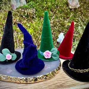 May include: Five miniature witch hats in different colors and styles. The hats are made of velvet and have various embellishments, including a pink rose, green leaves, and a gold braid.