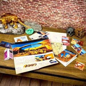 May include: A collection of miniature Arches National Park memorabilia including a gold-colored dinosaur figurine, a small skull, several printed brochures, postcards, and a Junior Ranger book spread across a wooden surface with a brick wall backdrop.