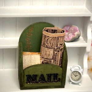 May include: A miniature green wooden mail holder with the word "MAIL" in black lettering. It contains rolled-up newspaper and aged paper. A small white clock and a pink and white stone are also visible.