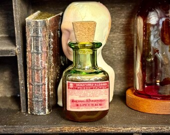 May include: A small, green glass bottle with a cork stopper and a red label reading "DENATURED ALCOHOL POISON CAUTION" sits on a shelf. The bottle is filled with a dark liquid. Other items on the shelf include a skull, a book, and rolled papers.