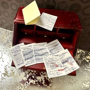 May include: A miniature wooden desk with a deep red finish, featuring small drawers and a hinged top. Several small papers with printed text are scattered around the desk, along with a folded yellow sticky note. The desk is set against a textured background.