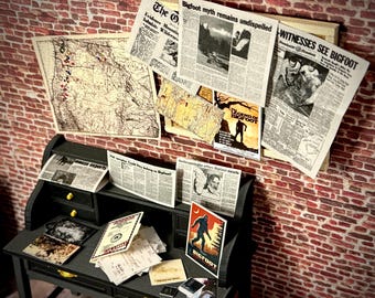 May include: A miniature black desk with drawers, topped with newspapers, maps, and a Bigfoot poster. Headlines include "Witnesses See Bigfoot" and "Bigfoot Myth Remains Undispelled." The background is a brick wall.