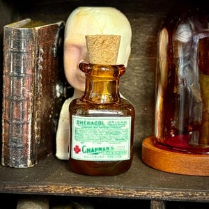 May include: An amber glass bottle with a cork stopper, labeled "Cheracol Poison" and "Chapman's" in green and white. The bottle is part of a collection of vintage apothecary items, including a book and a glass jar.
