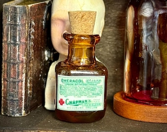 May include: An amber glass bottle with a cork stopper, labeled "Cheracol Poison" and "Chapman's" in green and white. The bottle is part of a collection of vintage apothecary items, including a book and a glass jar.