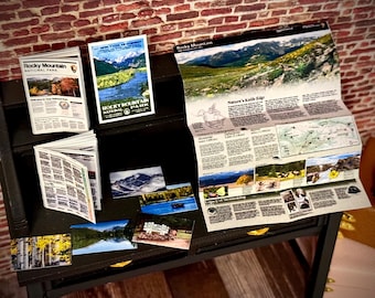 May include: A collection of miniature Rocky Mountain National Park souvenirs, including brochures, postcards, and a detailed map. The items are arranged on a small black table against a brick wall background. The postcards feature scenic views.