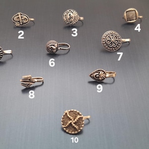 Puede incluir: Una colección de diez anillos de nariz plateados, cada uno con un diseño único. Los estilos incluyen patrones florales, geométricos y texturizados. Los anillos de nariz se muestran sobre una superficie gris oscuro.