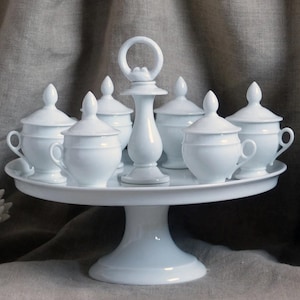 May include: A white porcelain serving set on a pedestal. The set includes a tiered cake stand with a handle, and six lidded cups with handles and saucers. The pieces are all white and have a classic design.
