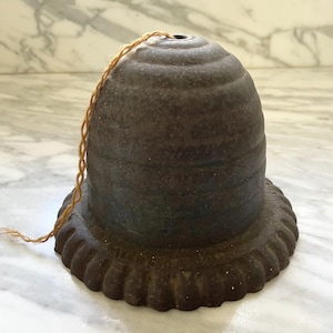 May include: A dark brown, antique-style lamp shade with a textured surface and a scalloped base. A twisted, tan-colored cord extends from the top. The shade has a bell-like shape and appears to be made of metal.