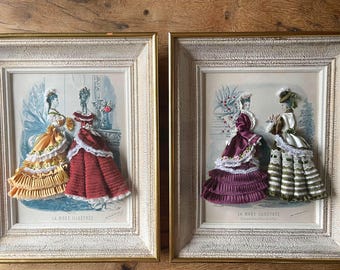 May include: Two framed vintage prints featuring detailed ribbon art of women in elegant dresses. The dresses are in shades of yellow, red, purple, and green, with white lace trim. The frames have a distressed, off-white finish and gold accents.