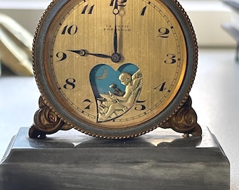 Puede incluir: Reloj antiguo dorado con esfera redonda y números romanos. La esfera tiene un recorte en forma de corazón con fondo azul y un ángel dorado. El reloj está sobre una base de mármol gris.