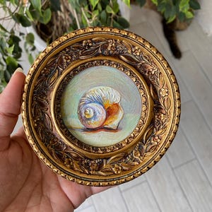 Könnte beinhalten: Ein kleines, gerahmtes Gemälde einer Muschel. Das Kunstwerk zeigt eine Muschel in Weiß-, Blau- und Orangetönen. Der Rahmen ist rund, goldfarben und mit detaillierten floralen und scrollartigen Mustern versehen. Das Gemälde wird in einer Hand gehalten.