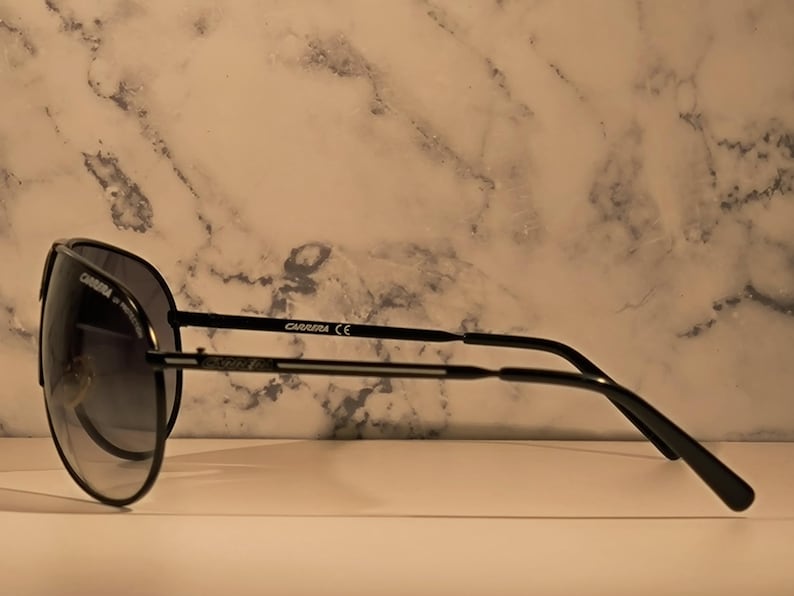 May include: Black Carrera sunglasses with a gradient lens. The sunglasses have a classic aviator shape with a thin black frame and the Carrera logo on the temple.