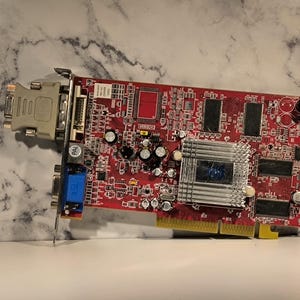 Pode incluir: Uma placa de circuito impresso vermelha com vários componentes eletrônicos, incluindo um dissipador de calor prateado, capacitores e conectores. A placa tem um conector de borda dourado e várias portas.