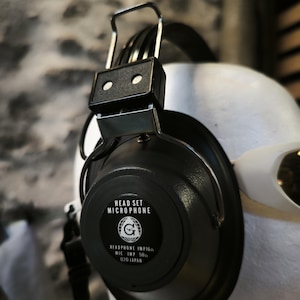 May include: Black over-ear headphones with a silver metal adjustable headband and a black circular earcup. The earcup features the text HEAD SET MICROPHONE, a globe logo with a G, and the text HEADPHONE IMP 16Ω MIC IMP 50Ω 020 JAPAN.