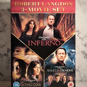 May include: A DVD box set titled "Robert Langdon 3-Movie Set" featuring Tom Hanks. The set includes the movies "Inferno", "Angels & Demons", and "The Da Vinci Code". The box has a red and black design with images from the films.