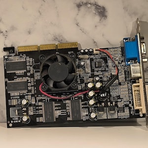 May include: A black computer graphics card with a cooling fan, various chips, and connectors. The card has a blue VGA port and a white DVI port. A red and black wire is connected to the fan.