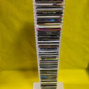 May include: A clear plastic CD tower filled with compact discs. The tower is rectangular with a white base. The CDs are stacked vertically, with some titles visible, including "The Very Best of Lou Reed" and "The Best of Roxy Music".
