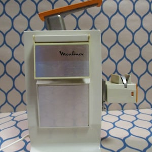 Puede incluir: Un abrelatas eléctrico Moulinex blanco con un mango naranja. El abrelatas tiene una cuchilla plateada y una base blanca.