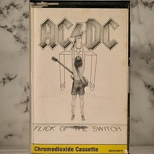 May include: A black and white cassette tape with a black and white image of a person flipping a switch. The cassette tape is labeled "AC/DC Flick of the Switch" and "Chromedioxide Cassette".