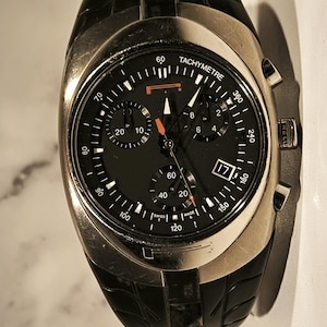 May include: A black and silver wristwatch with a black dial and a date display at the 3 o'clock position. The watch features multiple sub-dials and a tachymeter scale. The watch has a black leather strap.