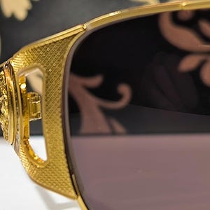 May include: Close-up of gold-colored sunglasses with a textured frame and dark lenses. The sunglasses have intricate detailing on the temples and a decorative design. The overall style is luxurious and fashionable.