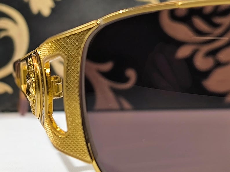 May include: Close-up of gold-colored sunglasses with a textured frame and dark lenses. The sunglasses have intricate detailing on the temples and a decorative design. The overall style is luxurious and fashionable.