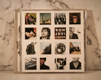 Puede incluir: Un álbum de CD "Crush" de Bon Jovi en una caja de plástico transparente. La portada del álbum presenta una cuadrícula de imágenes, incluyendo miembros de la banda, un primer plano de una persona y el título del álbum. El fondo tiene un patrón similar al mármol.